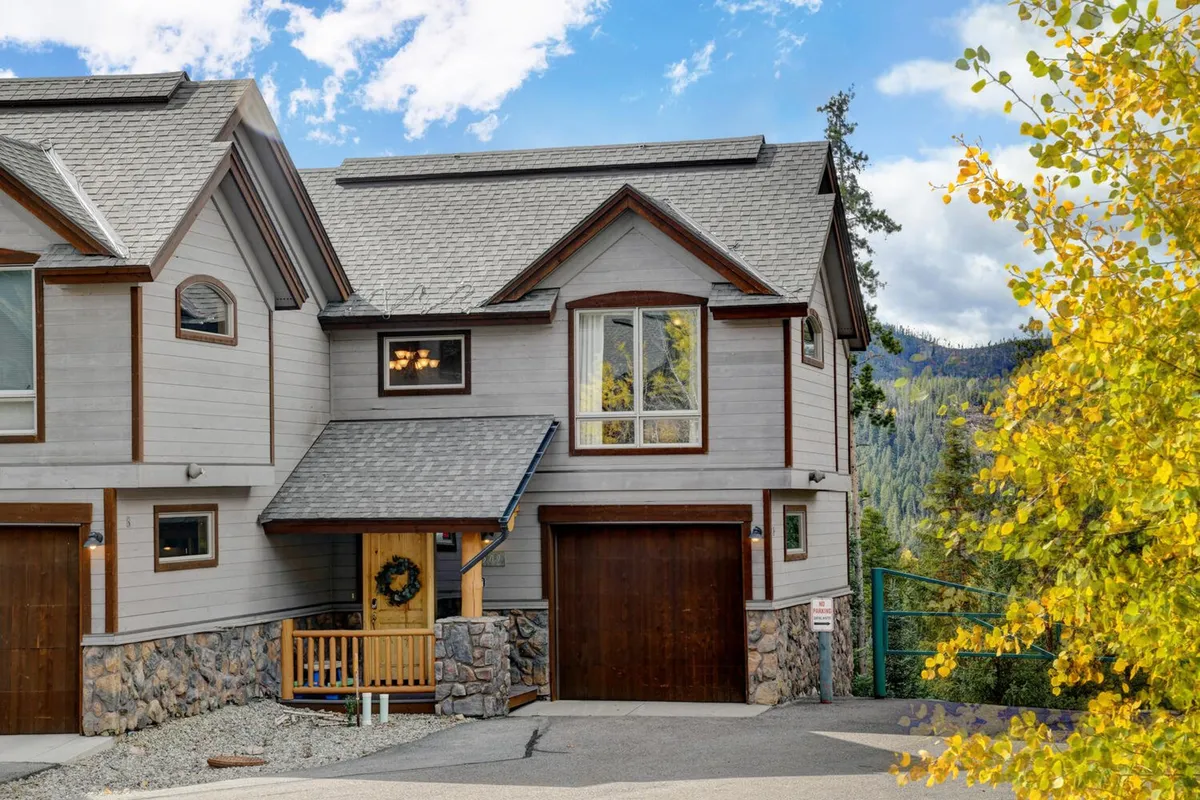 A welcoming exterior shot of the villa featuring the front entrance with a gabled roof, large window, stone foundation, and mature trees surrounding the property. The scene evokes the idea of gathering for family events or simply relaxing outdoors while enjoying the changing seasons.