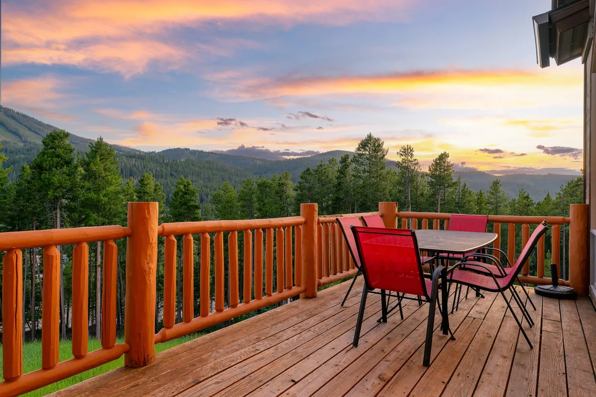 This exterior image captures a tranquil deck with a wooden railing, red chairs, a table set for dining, and sweeping mountain views at sunset. It visually extends the living/dining space outdoors, inviting guests to savor meals and mountain air al fresco.