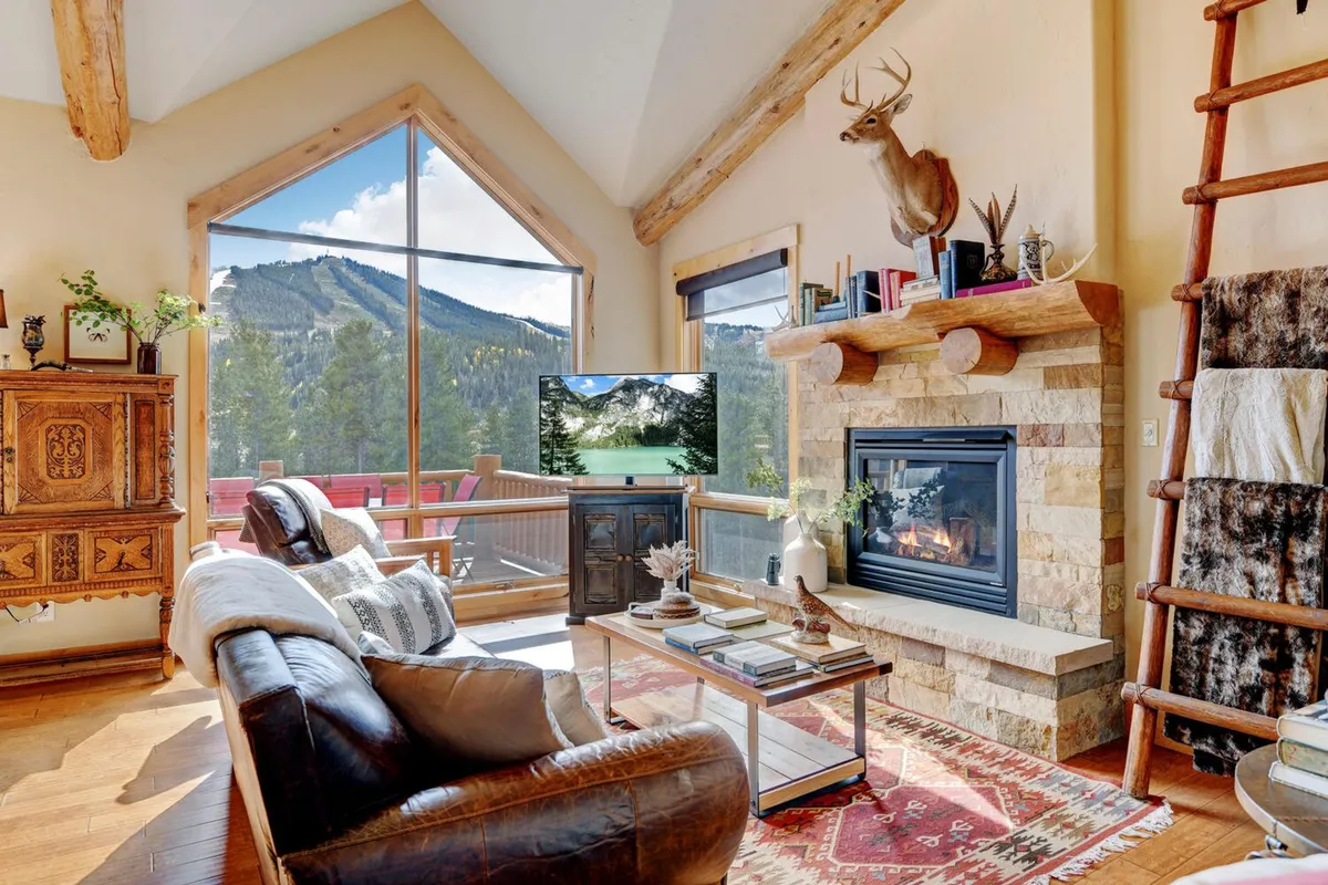 The image showcases a warm, inviting living room with rustic furnishings, a glowing fireplace, and a wall of windows framing breathtaking mountain views. Decorative antlers and a coffee table with books add to the cozy, alpine atmosphere, while natural light pours in to create a relaxing, social space perfect for unwinding or gathering with friends and family.