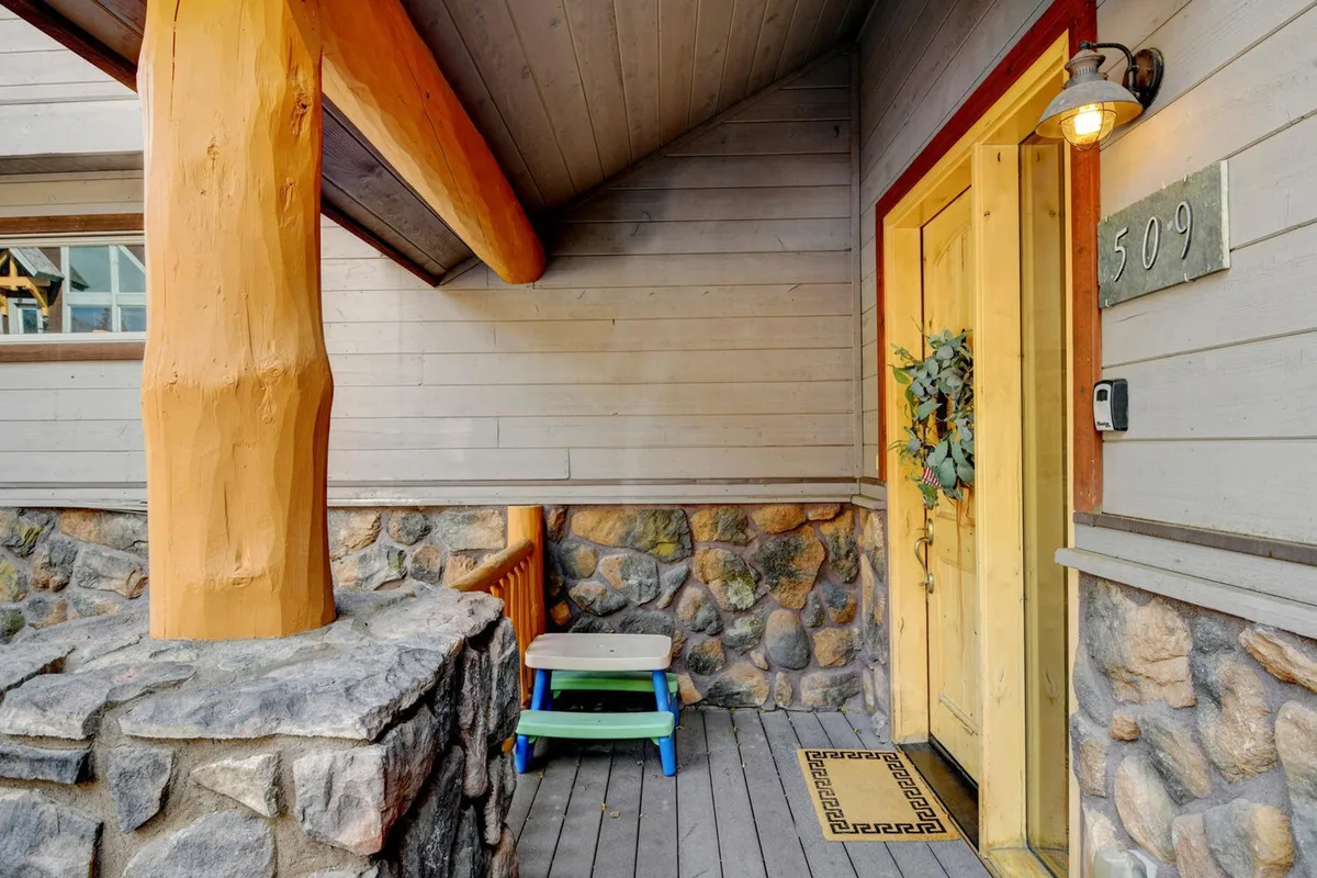 A warmly inviting exterior shot of the home's wooden front door, framed by stone steps, decorative wreath, and sturdy wooden beams. The entrance radiates cleanliness, hospitality, and first impressions of a well-kept, private residence.