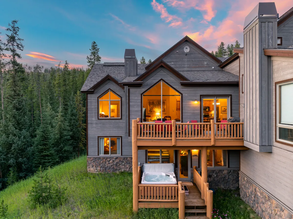 An expansive outdoor deck with a private hot tub, large windows reflecting the surrounding forest, and a serene sunset ambiance. The hot tub is ready for relaxation after a day on the slopes, and the space invites group gatherings and peaceful moments.
