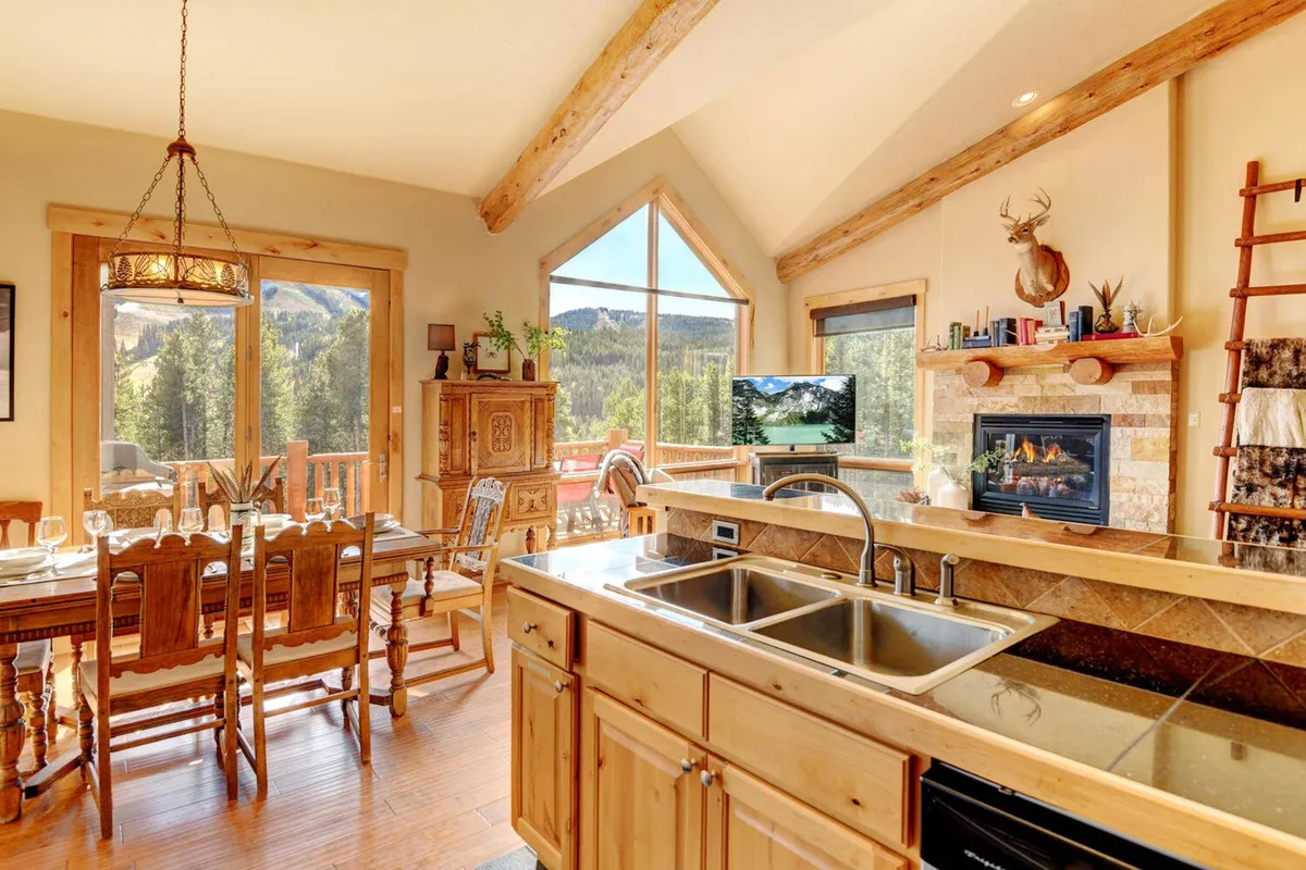 This image showcases a spacious open-concept area that seamlessly unites the dining table, kitchen island, stone fireplace, and large windows offering mountain views. The arrangement captures the social heart of the home, where family and friends can gather for meals, relax by the fire, and enjoy the scenery.
