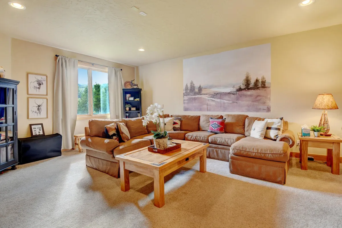 A spacious interior lounge featuring a large sectional sofa, a wooden coffee table, distinctive wall art, and a blue bookshelf. The layout suggests ample room for socializing, relaxing, or playing games, echoing the multi-purpose and family-friendly nature of the space.